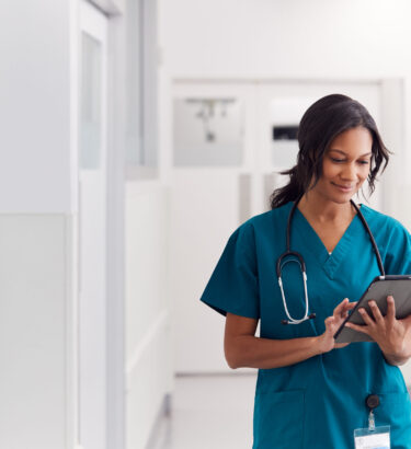 Female Doctor Wearing Scrubs In Hospital Corridor Using Digital Tablet Female Doctor Wearing Scrubs In Hospital Corridor Using Digital Tablet